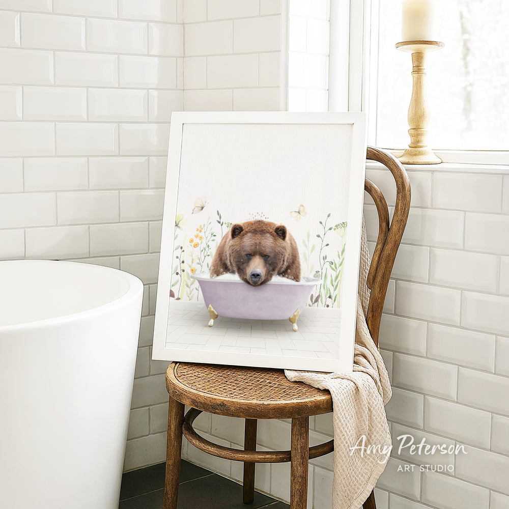 A framed print of a brown bear sitting in a bathtub is displayed on a wooden chair in a bathroom setting.