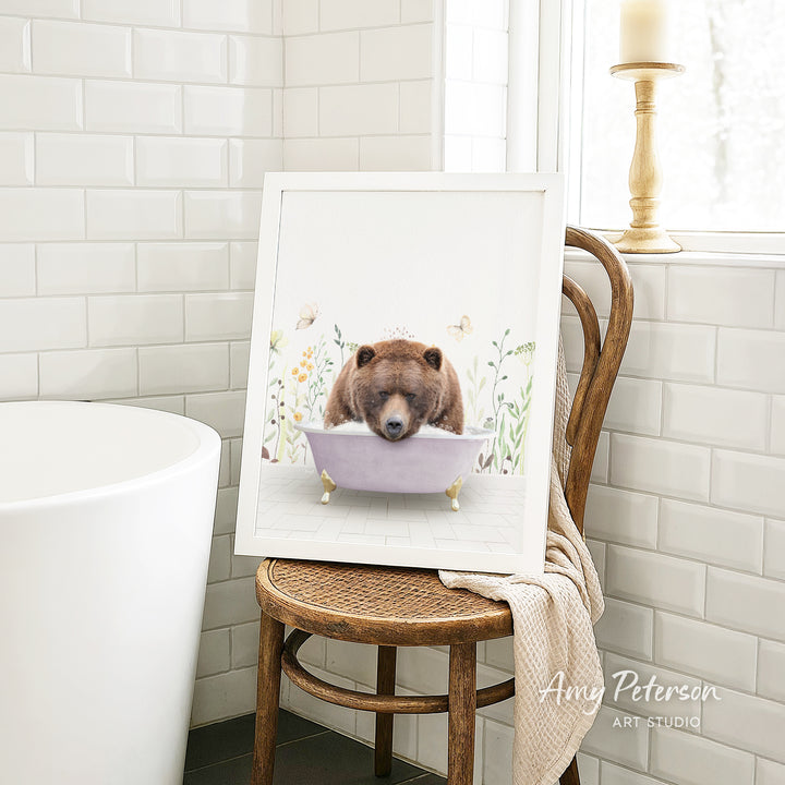 A framed print of a brown bear sitting in a bathtub is displayed on a wooden chair in a bathroom setting.