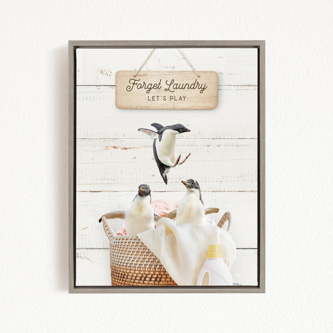 A framed image depicting three penguins in a basket, with a sign above them that says "Forget Laundry, Let's Play".