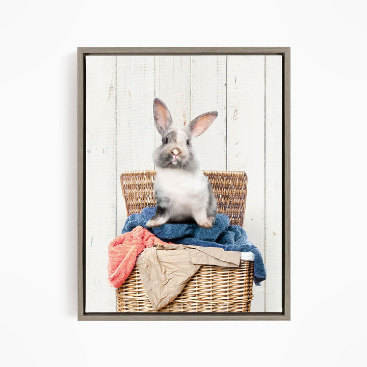 A gray and white rabbit sits in a wicker basket filled with clothes, against a white wooden background.