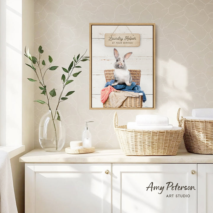 a framed picture of a rabbit sitting in a wicker basket, with a sign above it that reads "Laundry Helper at your service". The scene is set in a bathroom, with a white cabinet and a vase of greenery on the counter.