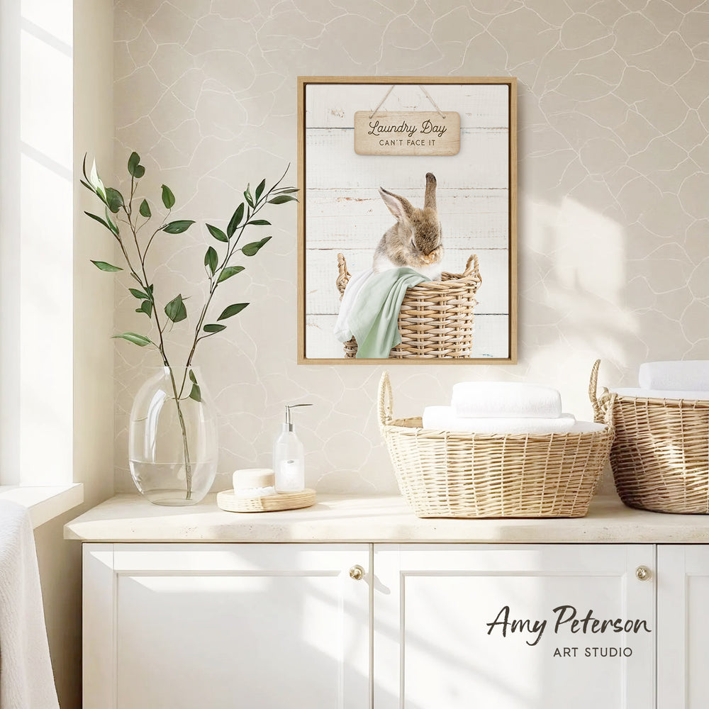 a framed picture of a rabbit sitting in a wicker basket, with a sign above it that says "Laundry Day Can't Face It". The scene is set in a bathroom, with a wicker basket, a soap dispenser, and a plant on the counter.