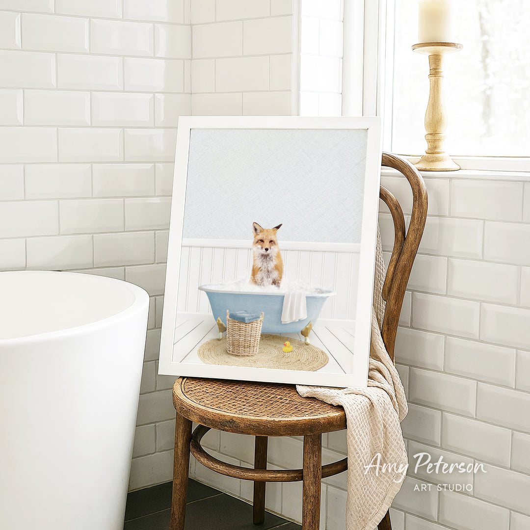 A framed image of a cat sitting in a bathtub, surrounded by a basket of bath toys, is displayed on a wooden chair in a bathroom setting.