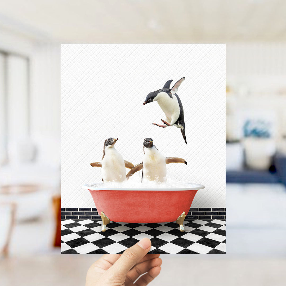 A hand holds up a framed image depicting two penguins sitting in a red bathtub, with one penguin flying out of the tub.