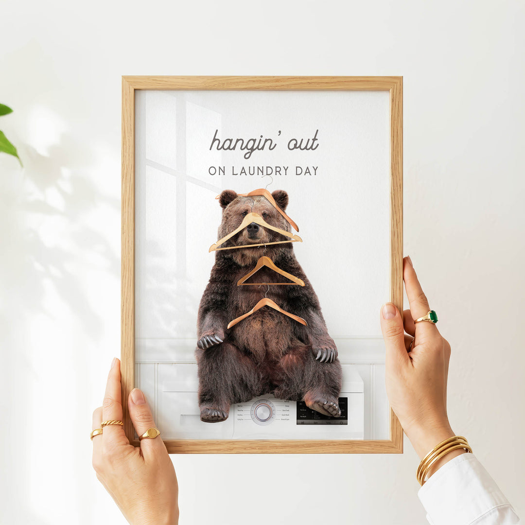 A person's hands are holding a framed picture of a bear sitting on a washing machine, with the bear holding multiple clothes hangers.
