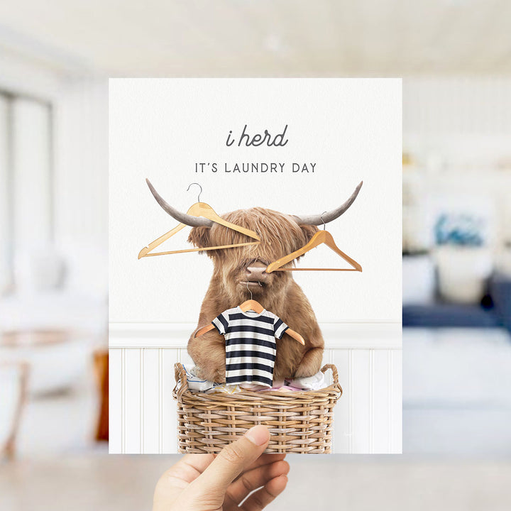 A hand holding a greeting card featuring a cartoon image of a Highland cow with clothes hanging from its horns, accompanied by the text "It's laundry day".