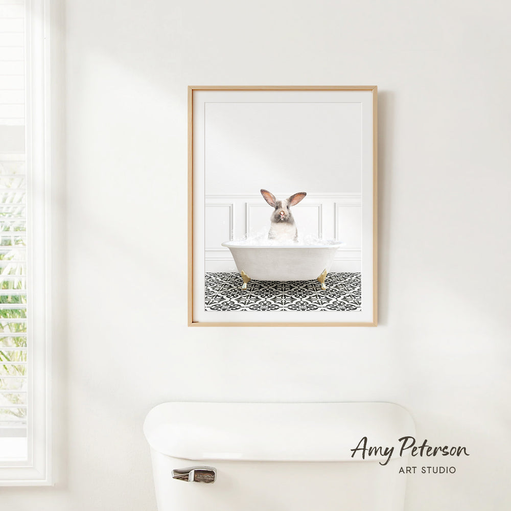 A framed image of a rabbit sitting in a bathtub, with a patterned tile floor beneath it, is hanging on a white wall.