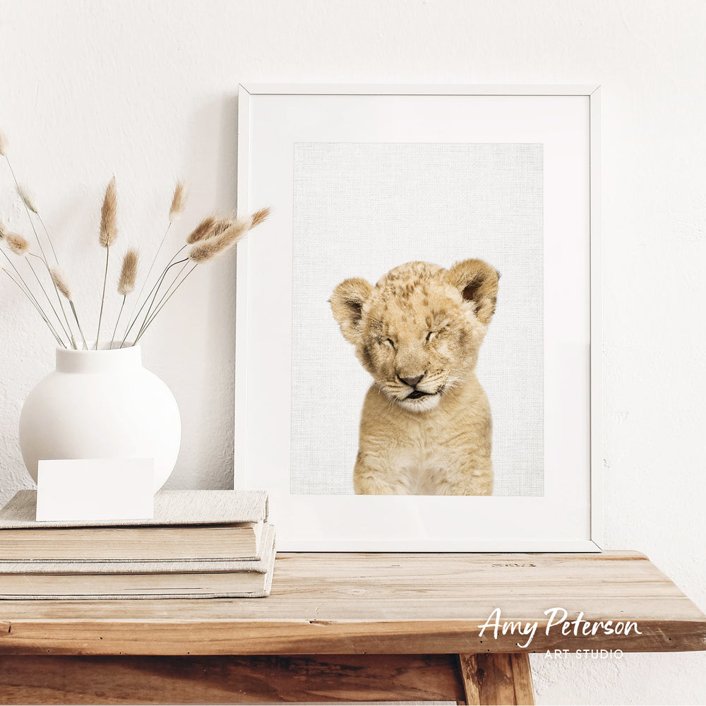 a young lion cub with its eyes closed, displayed in a white frame on a wooden shelf.