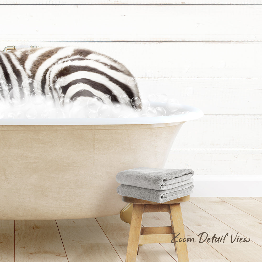 A zebra standing in a bathtub filled with bubbles, with a towel and stool nearby.
