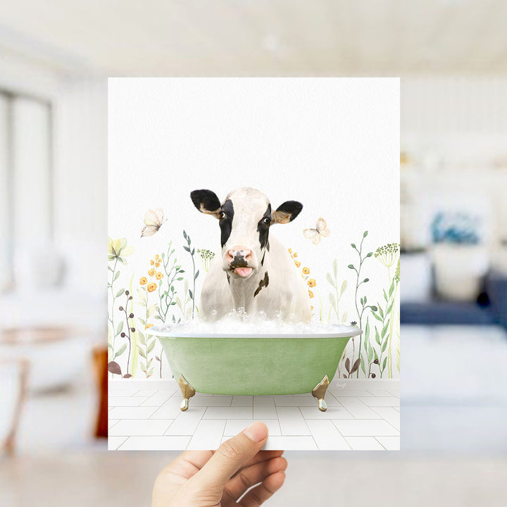 A hand holds up a framed image of a cow sitting in a green bathtub, surrounded by flowers and butterflies.