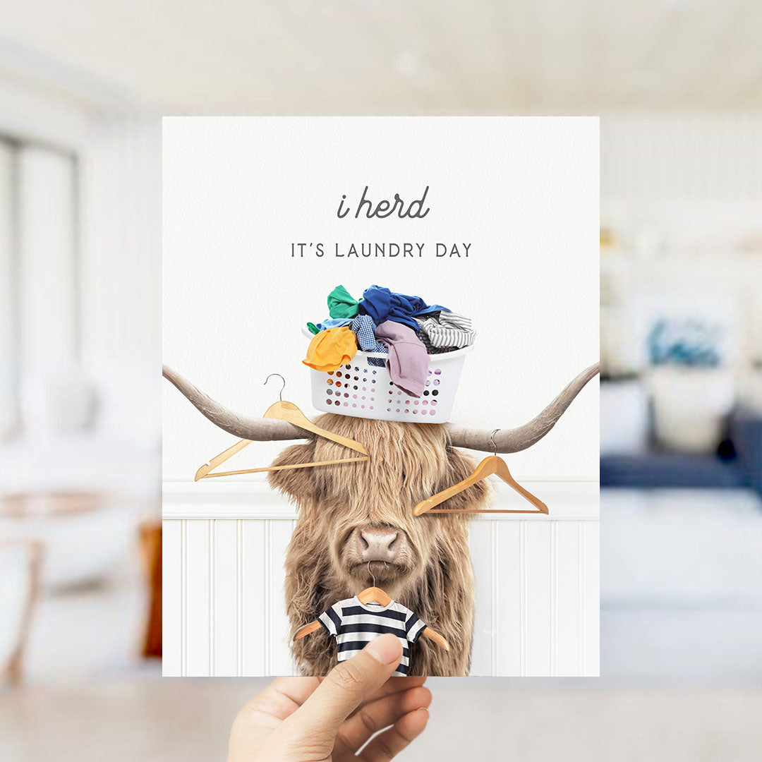 A hand holding a card with a picture of a cow wearing clothes and a basket of laundry on its head.