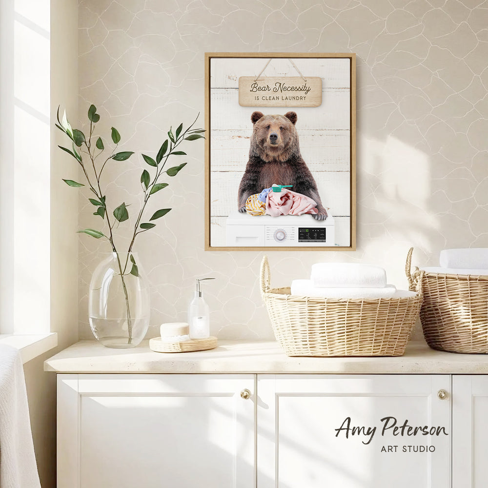 a framed poster of a bear sitting on a washer, with the text "Bear Necessity Is Clear Laundry" displayed above it. The poster is hanging on a wall above a white cabinet, which has a wicker basket and a soap dispenser on it. There is also a plant in a clear vase on the countertop.