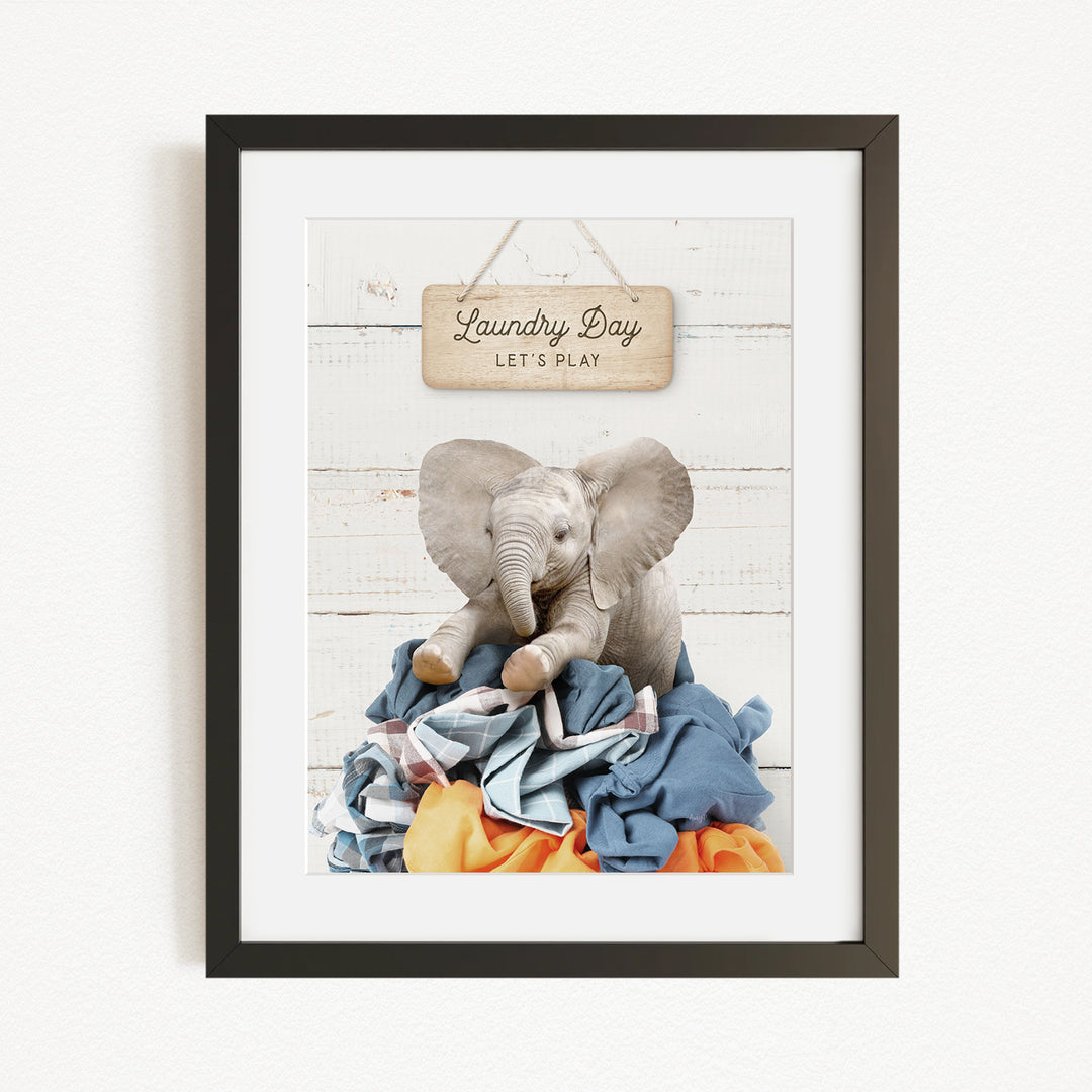 A framed image of a stuffed elephant sitting atop a pile of laundry, with a wooden sign above that says "Laundry Day Let's Play".