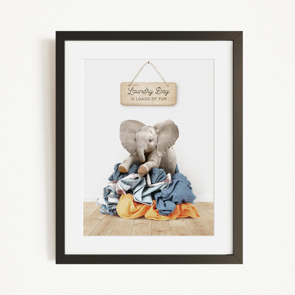 A framed image of a stuffed elephant sitting on a pile of laundry, with a sign above it that says "Laundry Day is loads of fun".