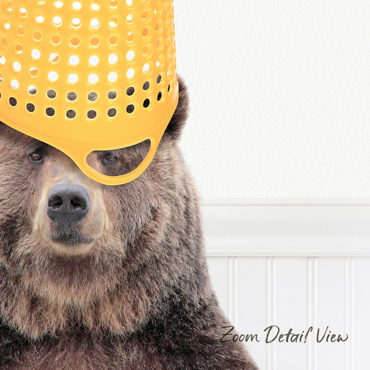 A close-up of a brown bear wearing a yellow plastic bucket hat, with a white wall in the background.
