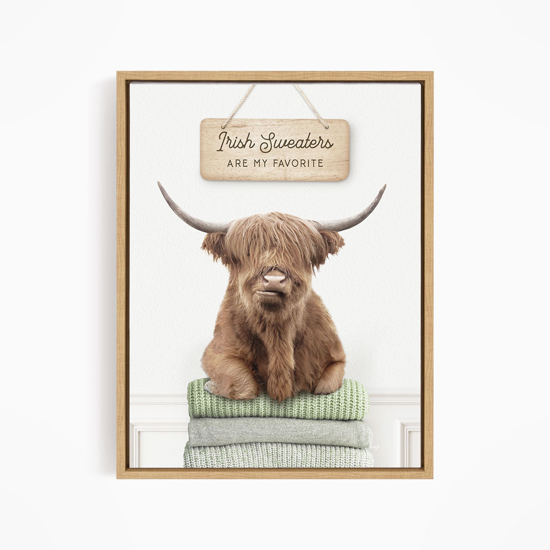 A brown Highland cow with large horns is sitting on a stack of green and gray blankets, with a wooden sign above that says "Irish Sweaters are my favorite".