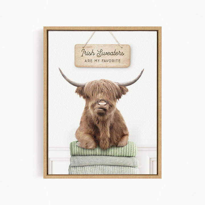 A brown Highland cow with large horns is sitting on a stack of green and gray blankets, with a wooden sign above that says "Irish Sweaters are my favorite".