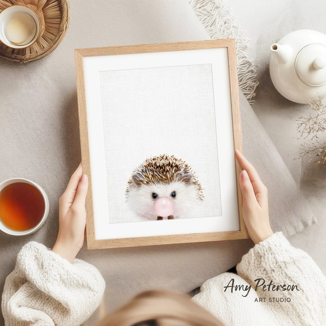 A person is holding a framed picture of a hedgehog with a pink bubble in its mouth, surrounded by various objects such as a teapot, a cup, and a bowl.