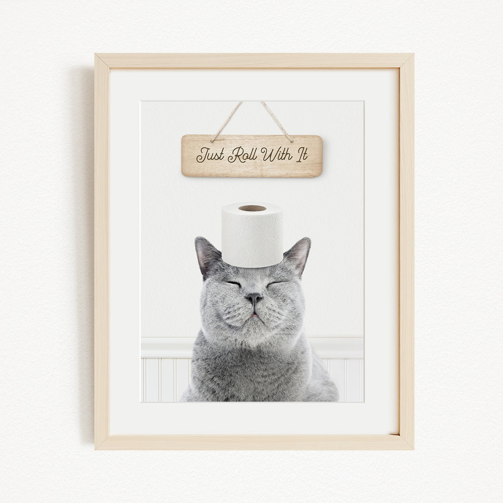 A gray cat sitting on a white surface with a wooden sign above it that says "Just Roll With It".