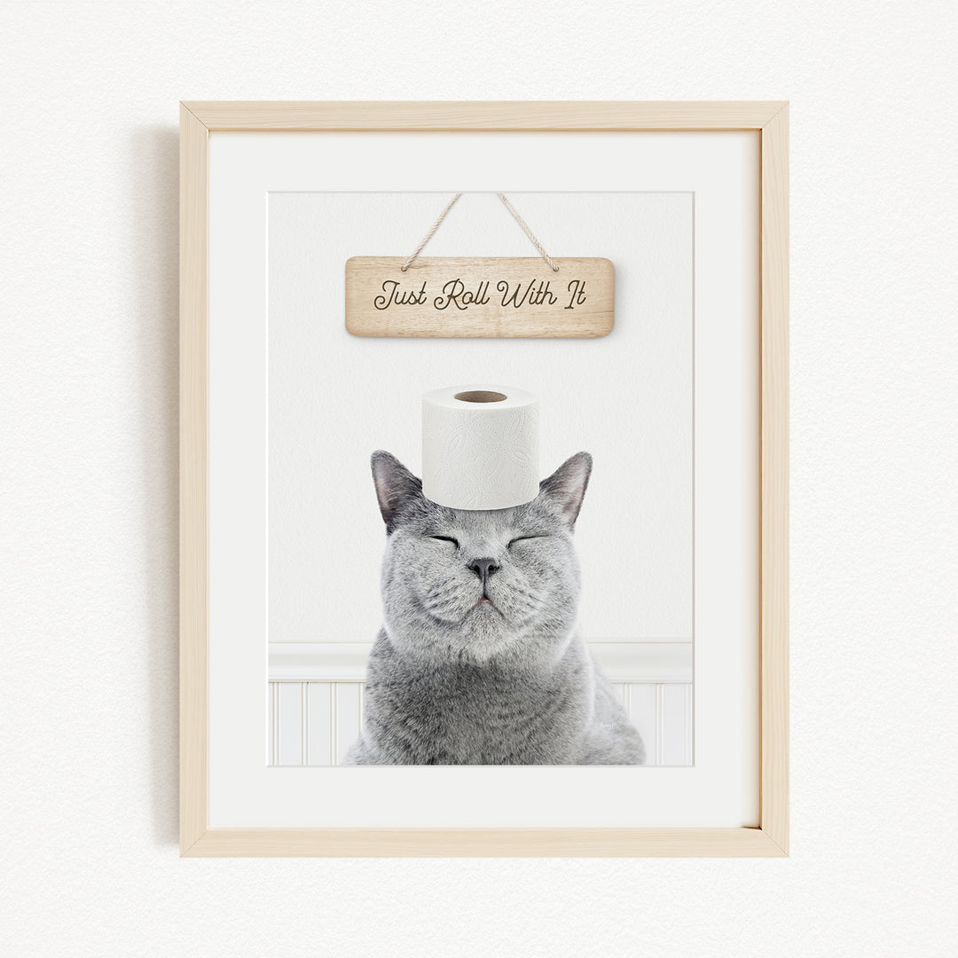 A gray cat sitting on a white surface with a wooden sign above it that says "Just Roll With It".