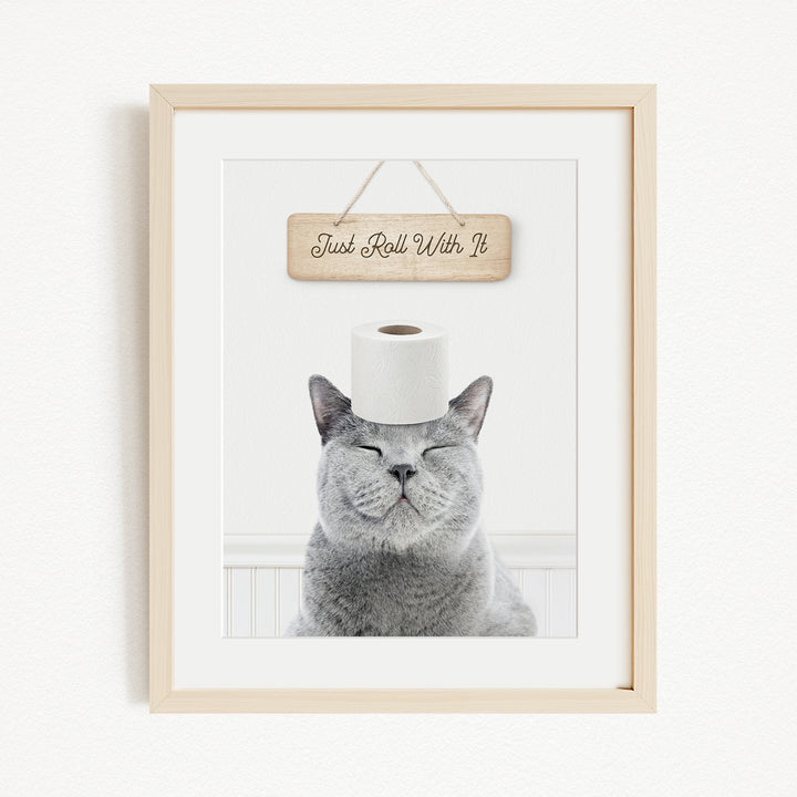 A gray cat sitting on a white surface with a wooden sign above it that says "Just Roll With It".
