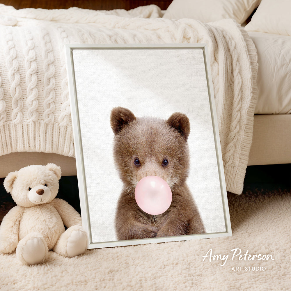 A framed image of a brown teddy bear blowing a bubble gum bubble, with a white teddy bear sitting next to it on a beige carpet.