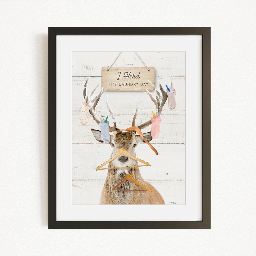 A framed print depicting a deer with antlers holding various laundry items, including socks and clothes hangers, with a sign above that says "I Harded It's Laundry Day".
