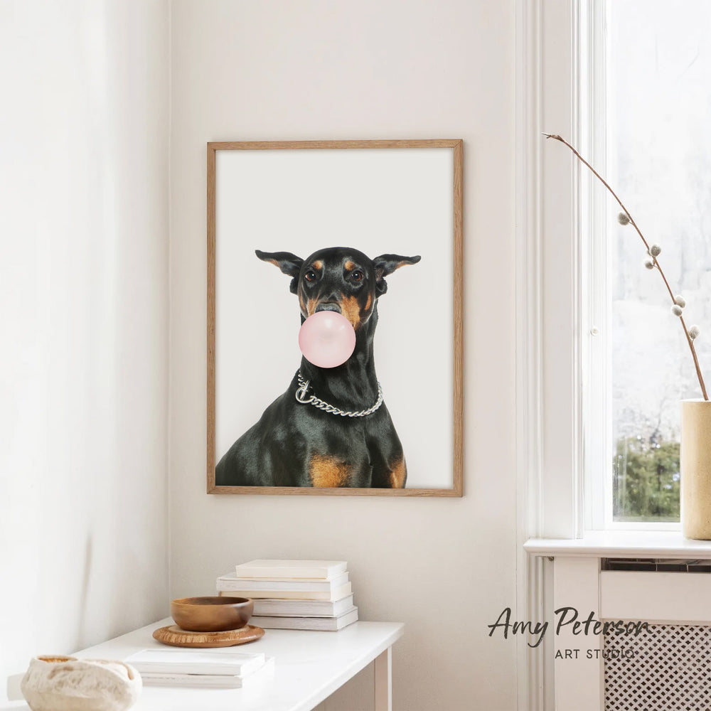 A framed image of a black and tan dog blowing a bubble with a pink bubble gum bubble, hanging on a white wall in a room with a white table and a window.