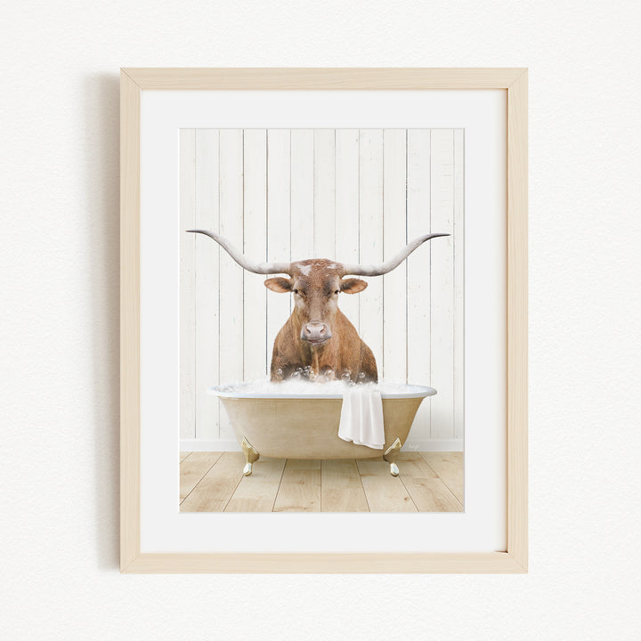 A framed image depicting a brown cow with large horns sitting in a white bathtub filled with water.