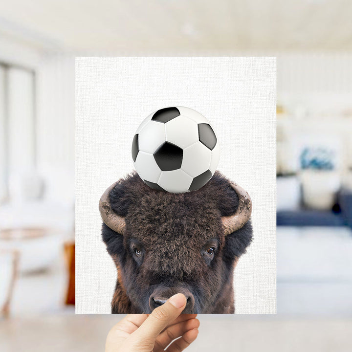 A hand holds up a picture of a bison with a soccer ball on its head.