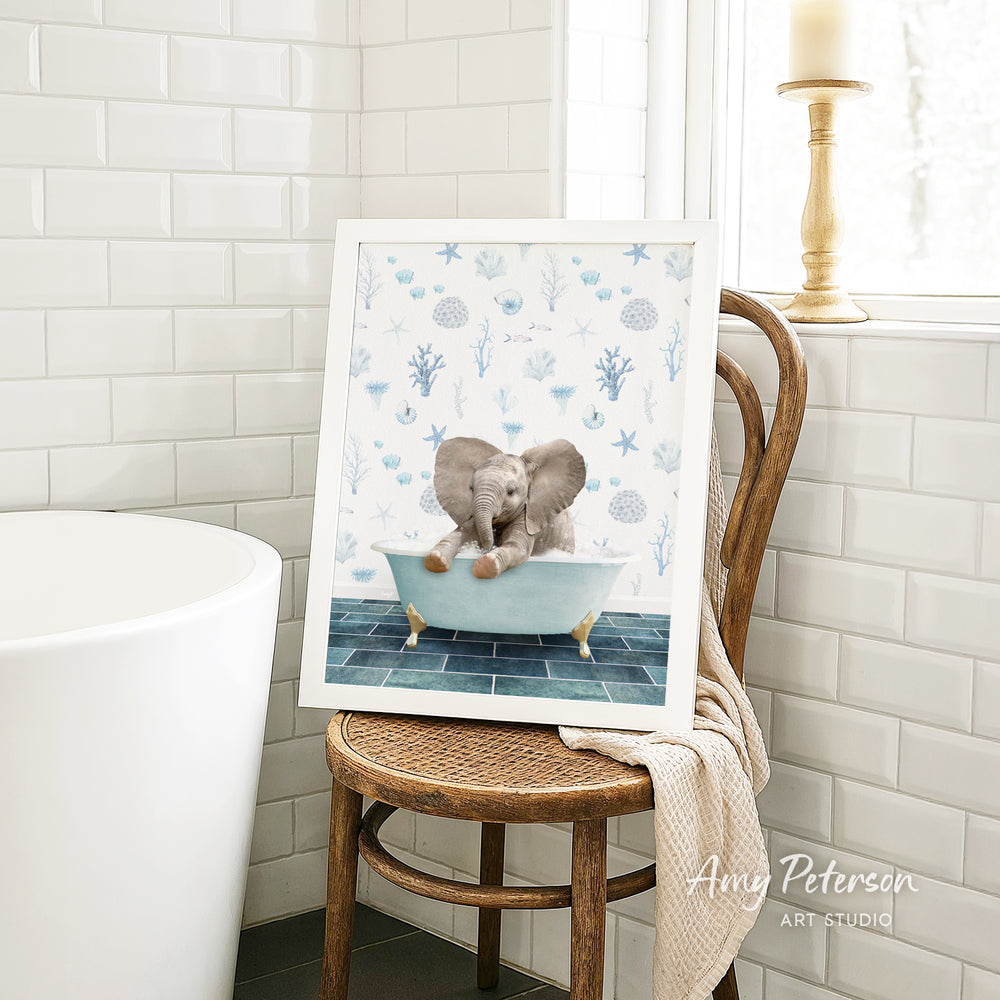 A framed image of a baby elephant sitting in a bathtub, surrounded by blue and white seashells and starfish, is displayed on a wooden chair in a bathroom setting.