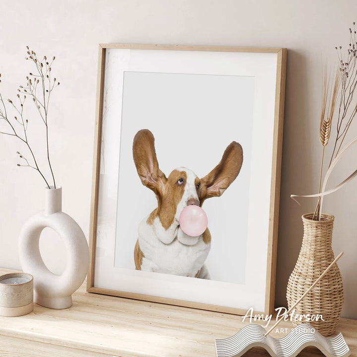 A framed photograph of a dog blowing a bubble gum bubble is displayed on a wooden shelf, accompanied by decorative vases and a woven basket.