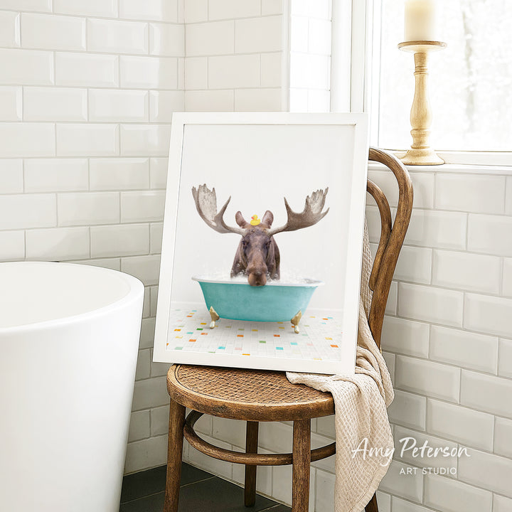 A framed image of a moose sitting in a blue bathtub, with its antlers raised, is displayed on a wooden chair in a bathroom setting.