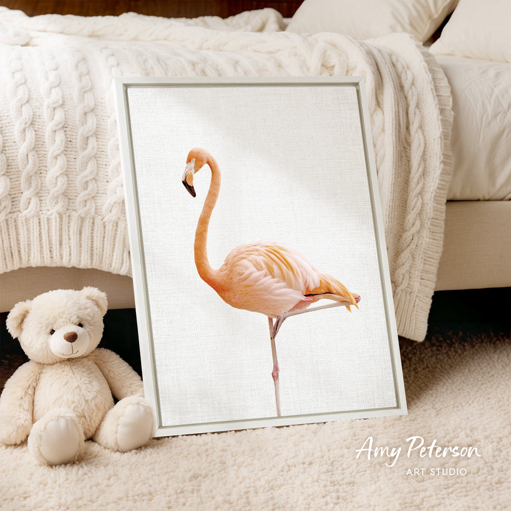 A framed image of a flamingo is displayed on a white surface, with a teddy bear sitting next to it.