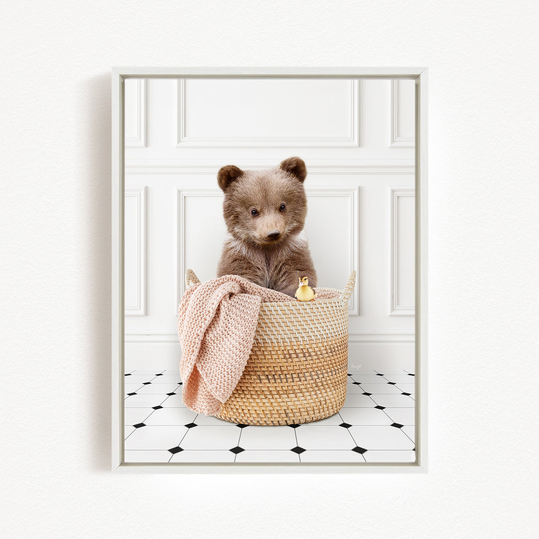 A brown bear sitting in a woven basket, with a pink blanket and a small yellow duck nearby, against a white wall with a black and white tiled floor.