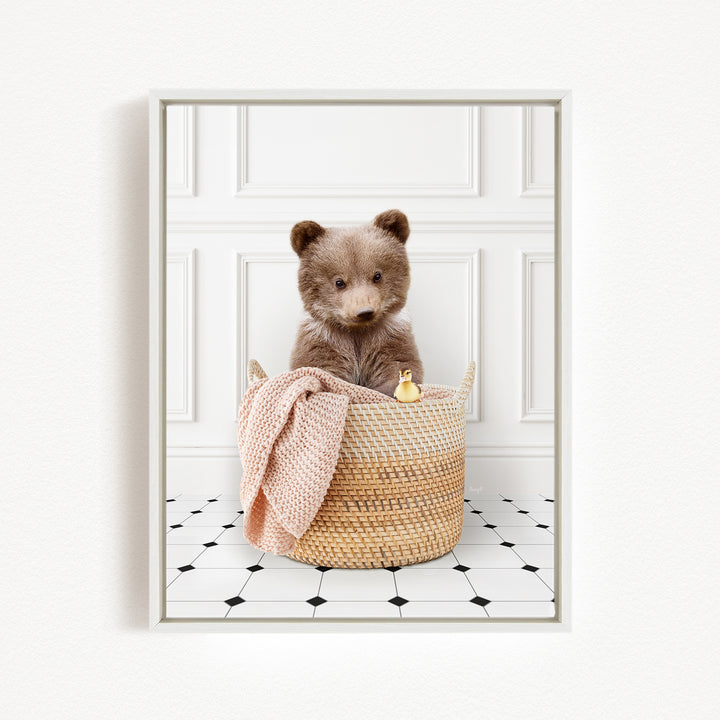 A brown bear sitting in a woven basket, with a pink blanket and a small yellow duck nearby, against a white wall with a black and white tiled floor.