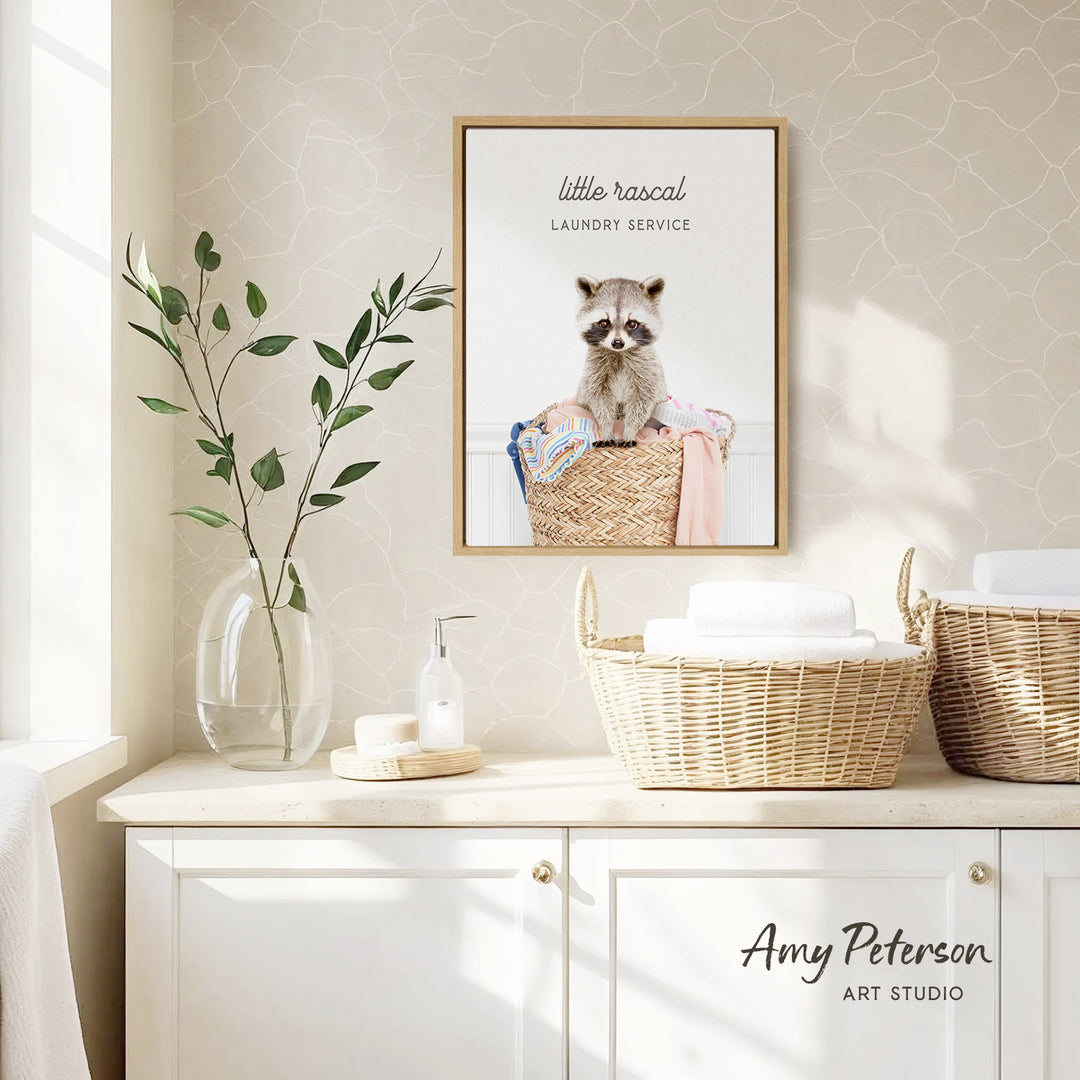 a framed photograph of a raccoon sitting in a laundry basket, with the text "Little raccoon laundry service" above it. The photograph is displayed on a wall above a white cabinet, which has a vase of leaves and a bottle of soap on it.