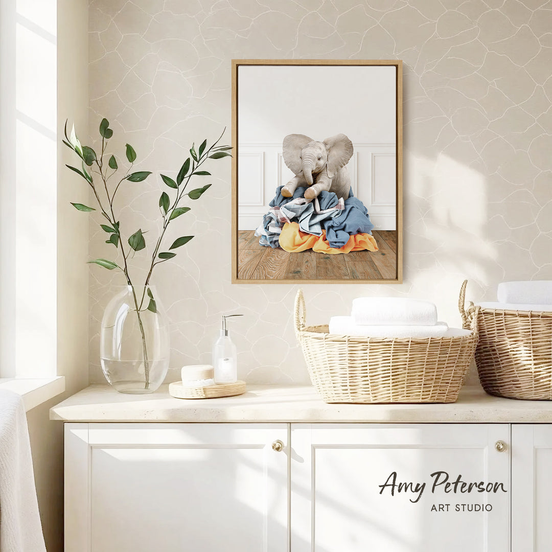 a bathroom scene with a framed photograph of two elephants sitting on a pile of clothes hanging on the wall. The photograph is framed in a white wooden frame and is placed above a white cabinet. The cabinet has two woven baskets on top, and there is a vase with green leaves on the counter. The wall behind the cabinet has a patterned wallpaper.