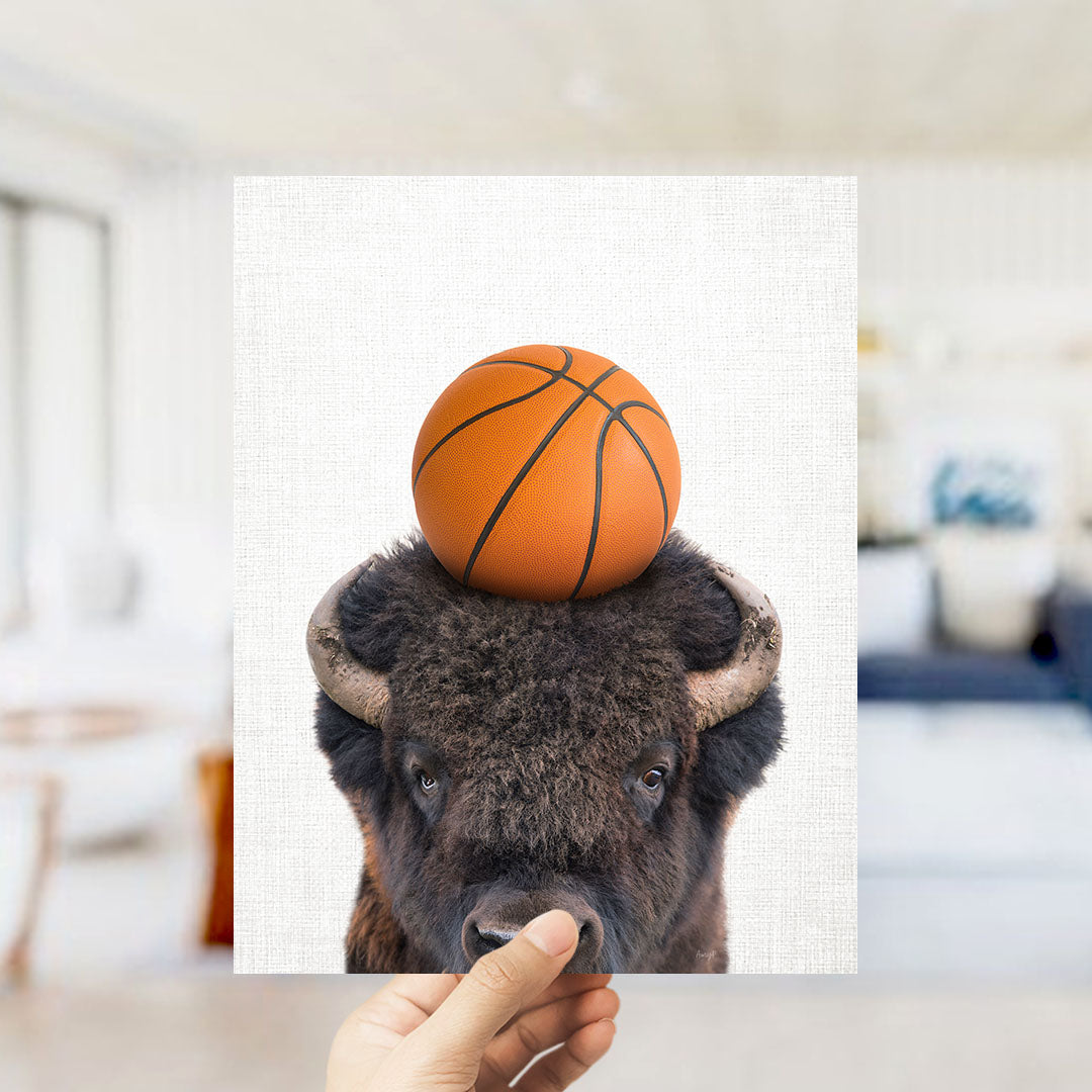 A hand holds up a picture of a buffalo with a basketball on its head.