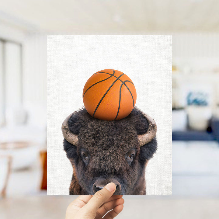 A hand holds up a picture of a buffalo with a basketball on its head.