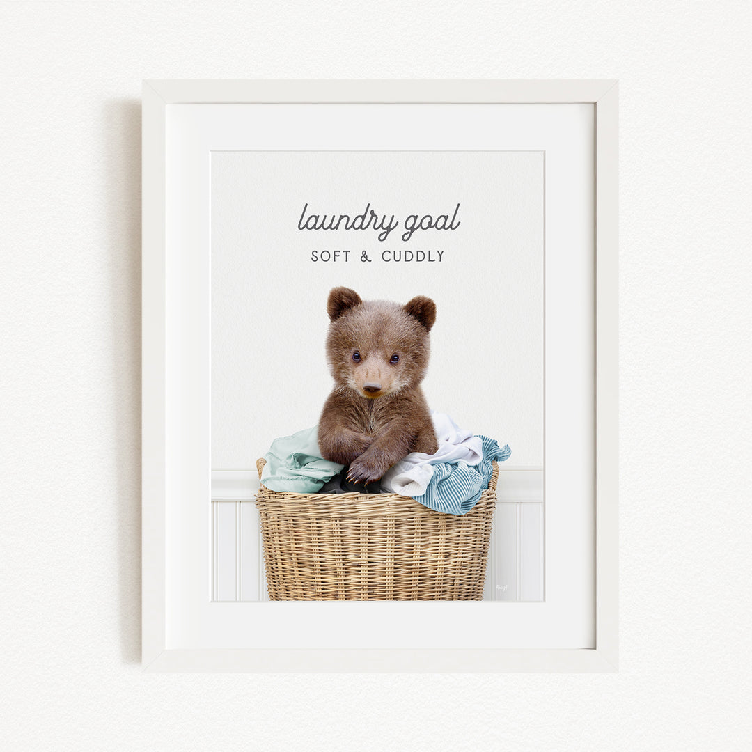 A cute brown bear sitting in a wicker laundry basket filled with clothes, with the text "laundry goal SOFT & CUDDLY" displayed above the bear.