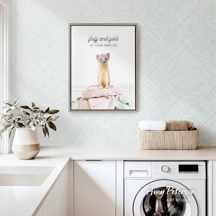 a framed poster of a ferret sitting on a pile of clothes, hanging on a wall above a laundry basket and a white sink.