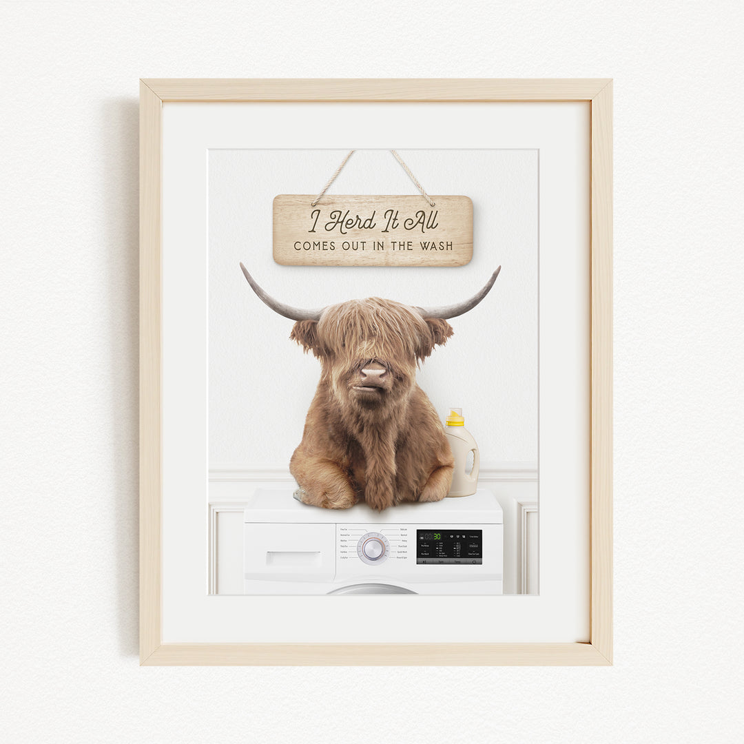 A framed image depicting a large, shaggy-haired animal, possibly a yak or a yak dog, sitting on top of a washing machine. The animal appears to be looking directly at the camera. The image also includes a sign that reads "I herd it all comes out in the wash".