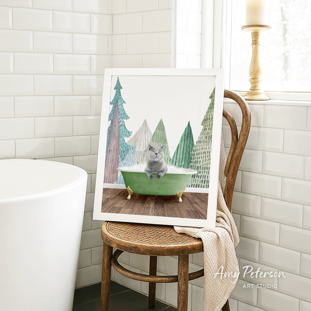 A framed image of a gray cat sitting in a green bathtub, surrounded by Christmas trees, is displayed on a wooden chair in a bathroom setting.