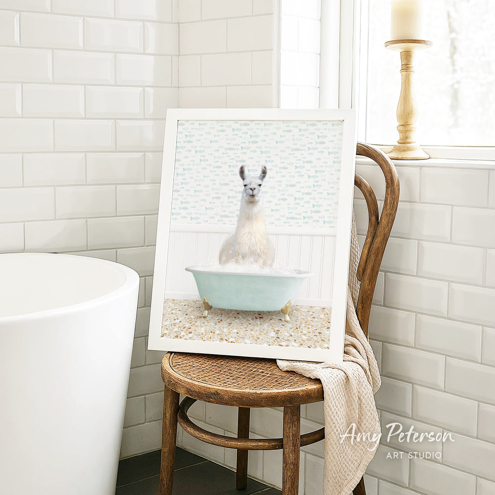 A framed image of a white llama sitting in a blue bathtub is displayed on a wooden chair in a bathroom setting.