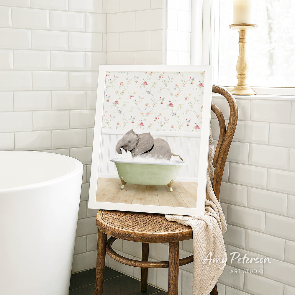 A framed image of a gray elephant sitting in a green bathtub, surrounded by a floral pattern, is displayed on a wooden chair in a bathroom setting.