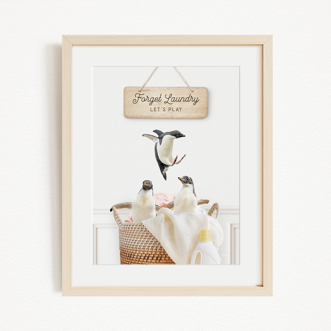 A framed image of three penguins in a basket, with a wooden sign above them that says "Forget Laundry, Let's Play".