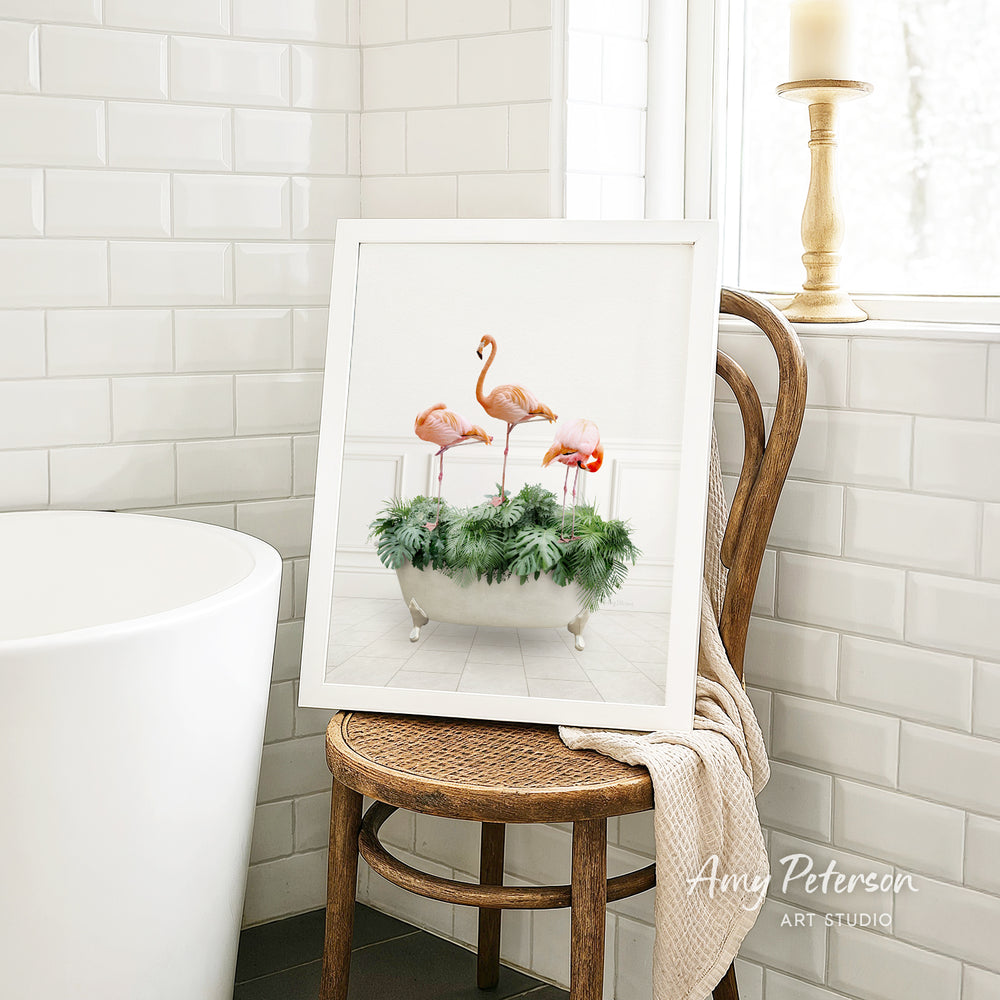 A framed image of three pink flamingos standing in a bathtub filled with green plants is displayed on a wooden chair in a bathroom setting.