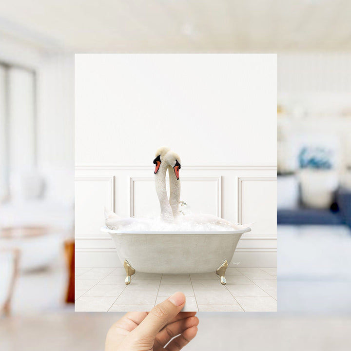 A hand holds up a framed photograph of two swans in a bathtub, with the swans appearing to be splashing and enjoying the water.