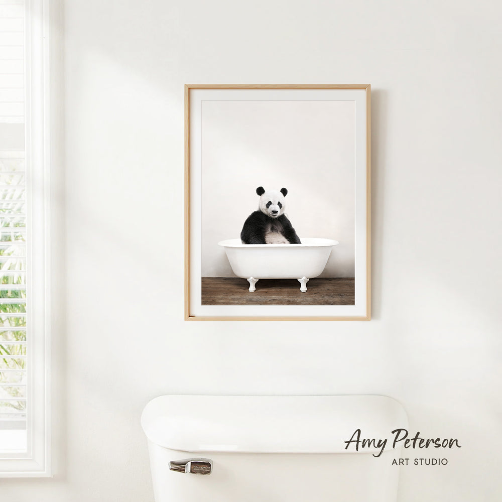 A framed photograph of a panda bear sitting in a bathtub is hanging on a white wall.
